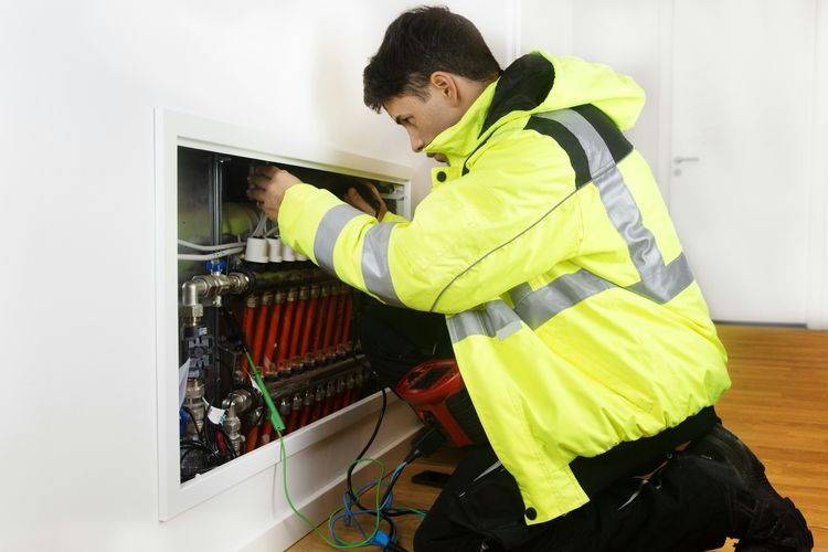 A plumber in a high-visibility jacket working on a water manifold in a wall cabinet